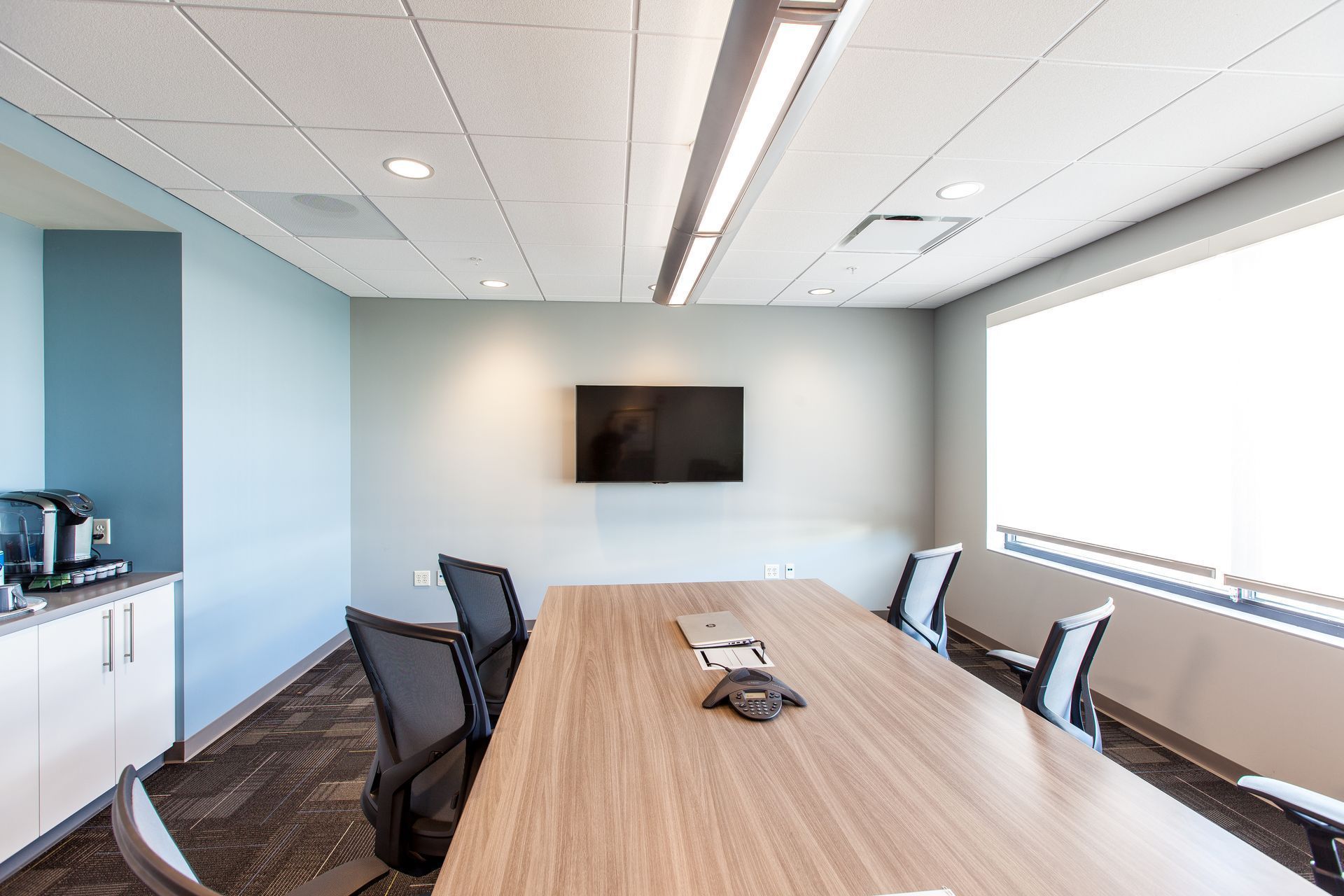 Modern conference room with a long wood table, black chairs, wall-mounted TV, and large bright windows