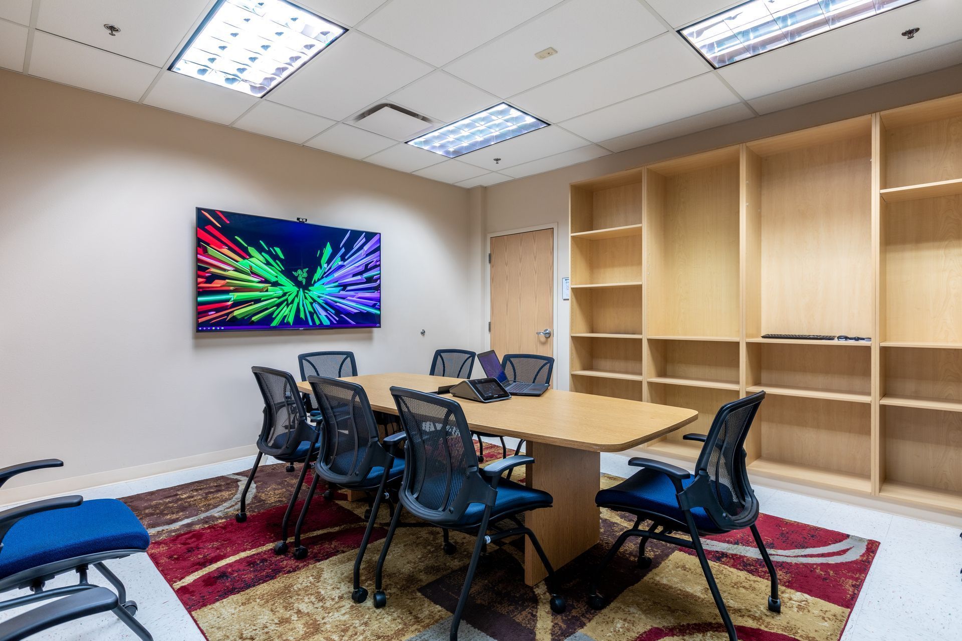 Conference room with table, black chairs, wall screen, and empty wooden shelves on patterned rug.