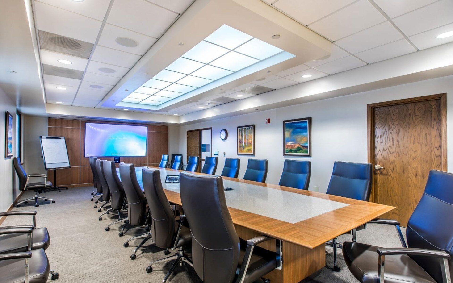 Conference room with long wooden table, blue chairs, wall art, and a presentation screen on the far wall