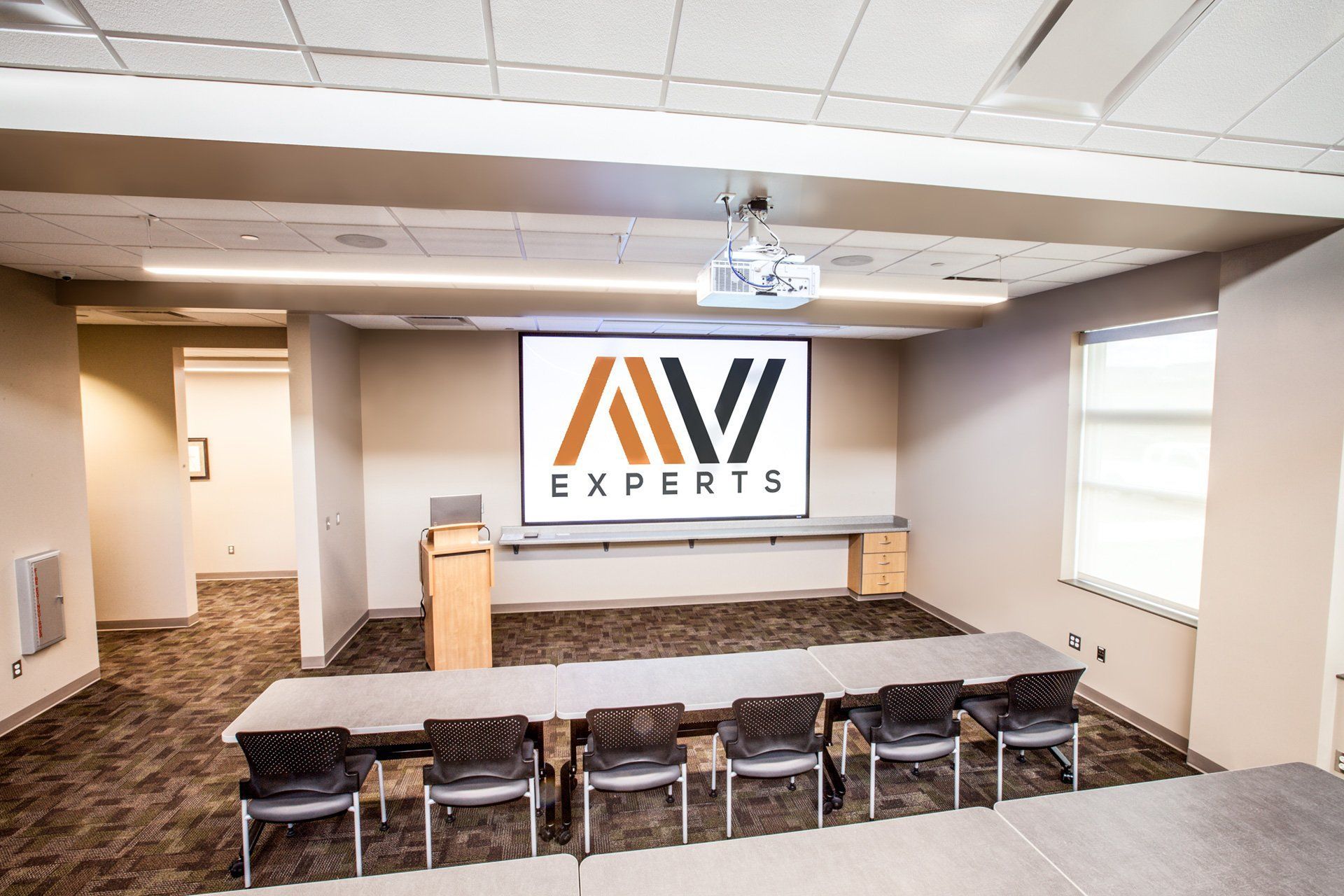 Conference room with AV Experts logo on the front wall, long table, chairs, and projector screen on the ceiling