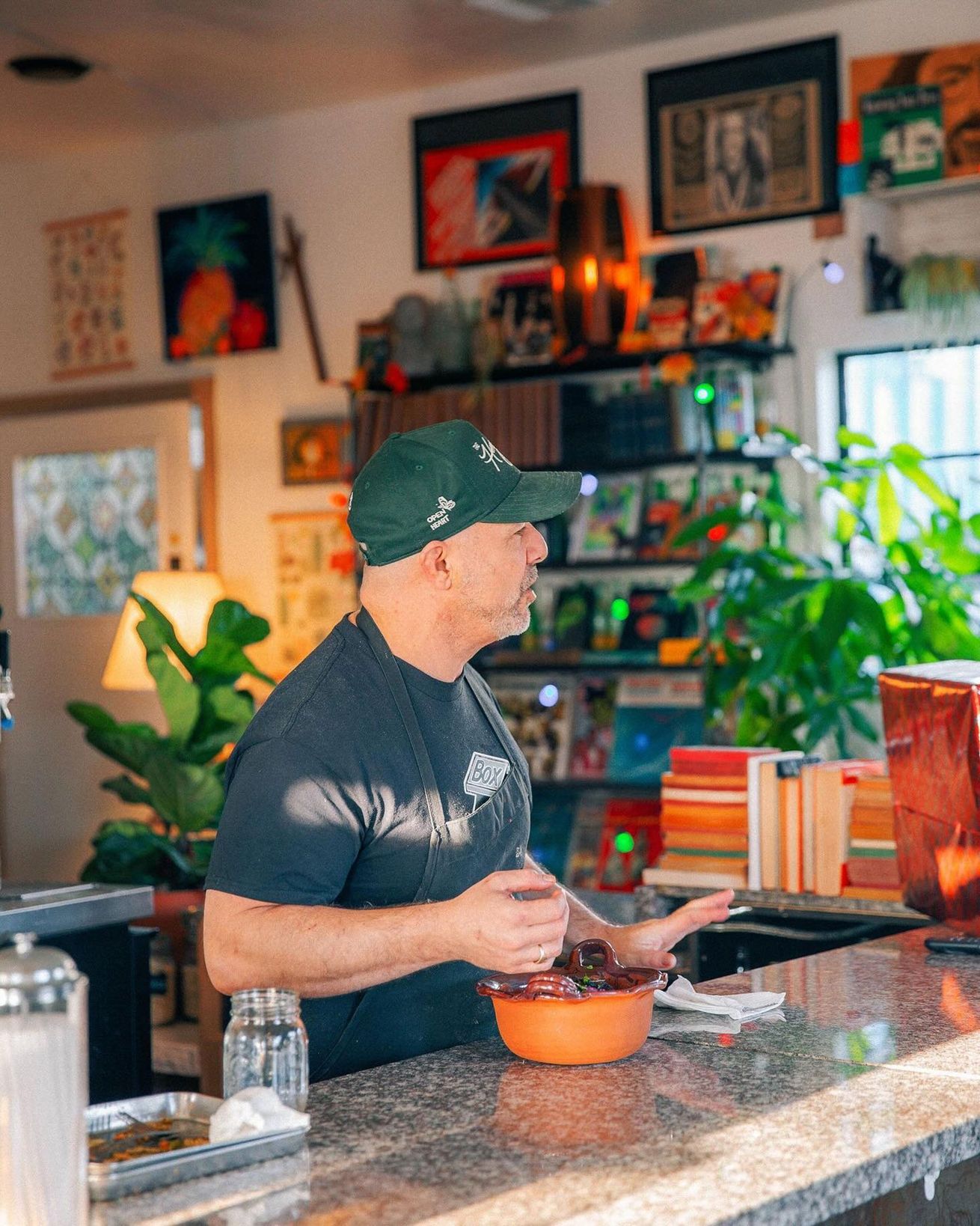 Man in green cap behind a counter, holding a bowl, talking. Shop interior with plants, artwork.