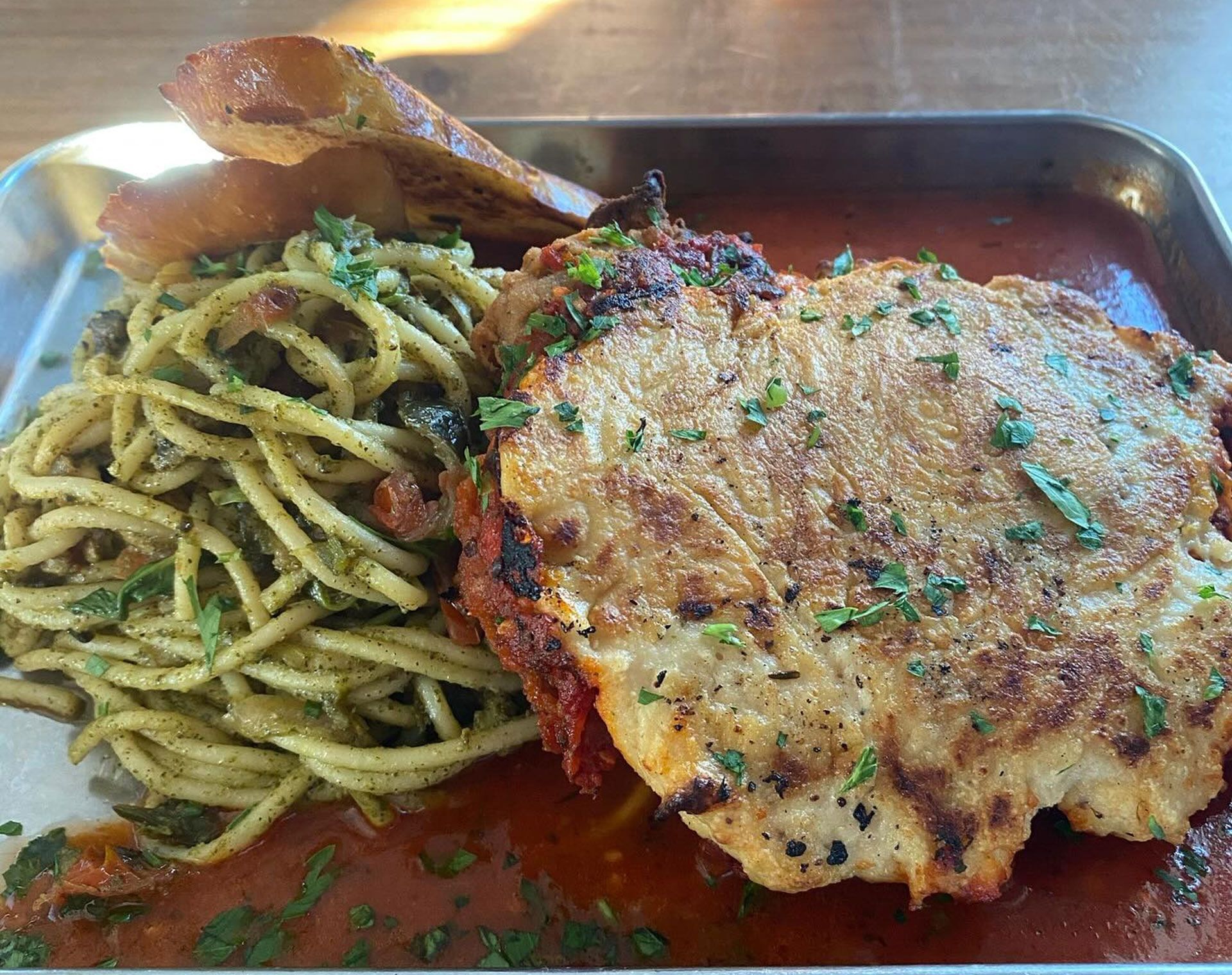 Chicken cutlet with spaghetti in pesto and garlic bread on a metal tray.
