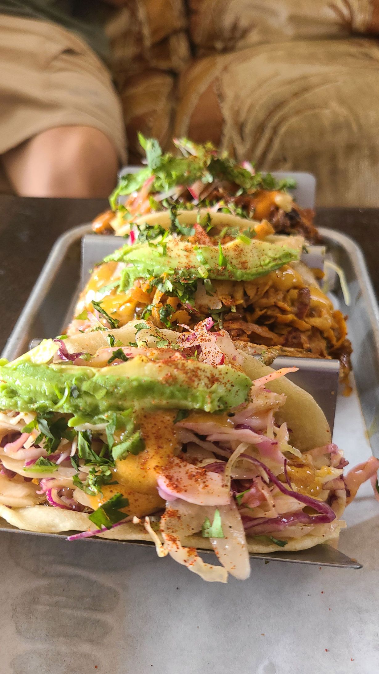 Three tacos on a tray, topped with avocado, slaw, and cilantro.