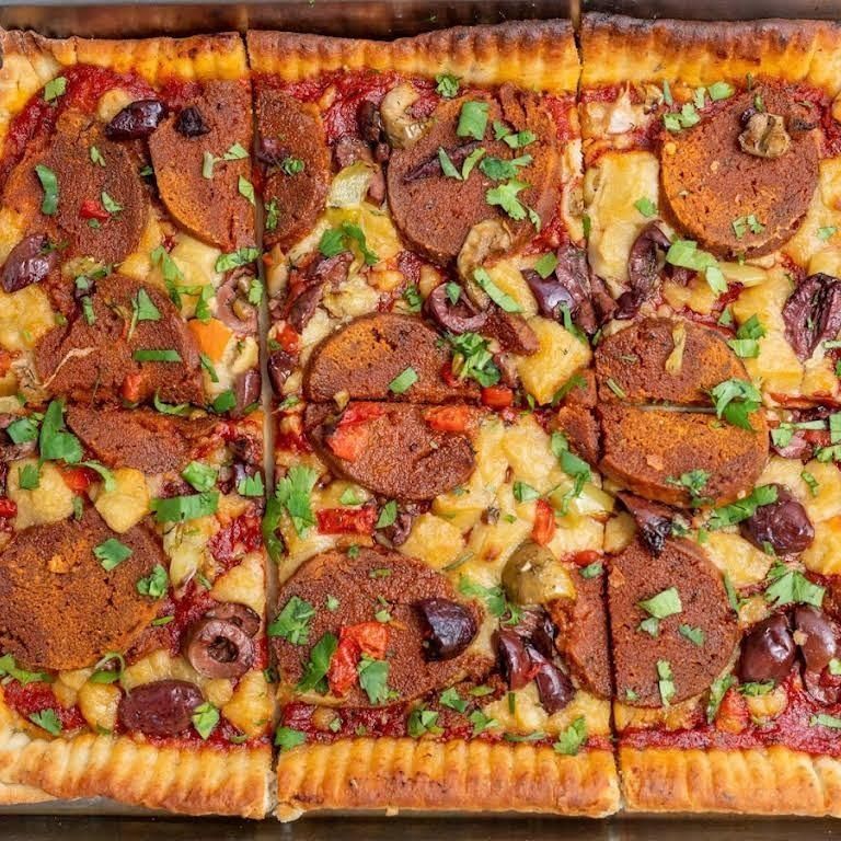Square pizza with pepperoni, olives, and herbs, cut into squares.