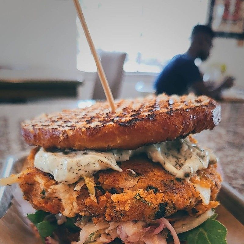 Burger with toasted bun, patty, sauce, and greens, on a tray. A person is blurred in the background.
