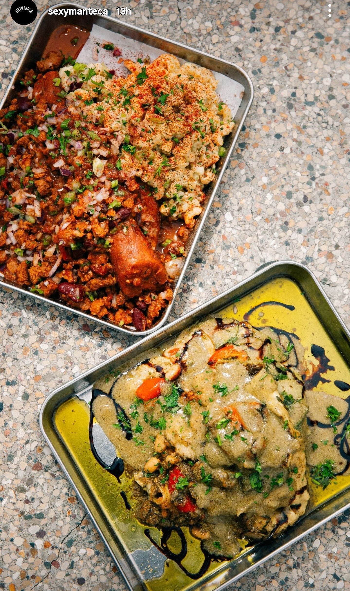 Two metal trays of food. One with a meat and grain dish. The other with a saucy dish drizzled with a darker sauce.