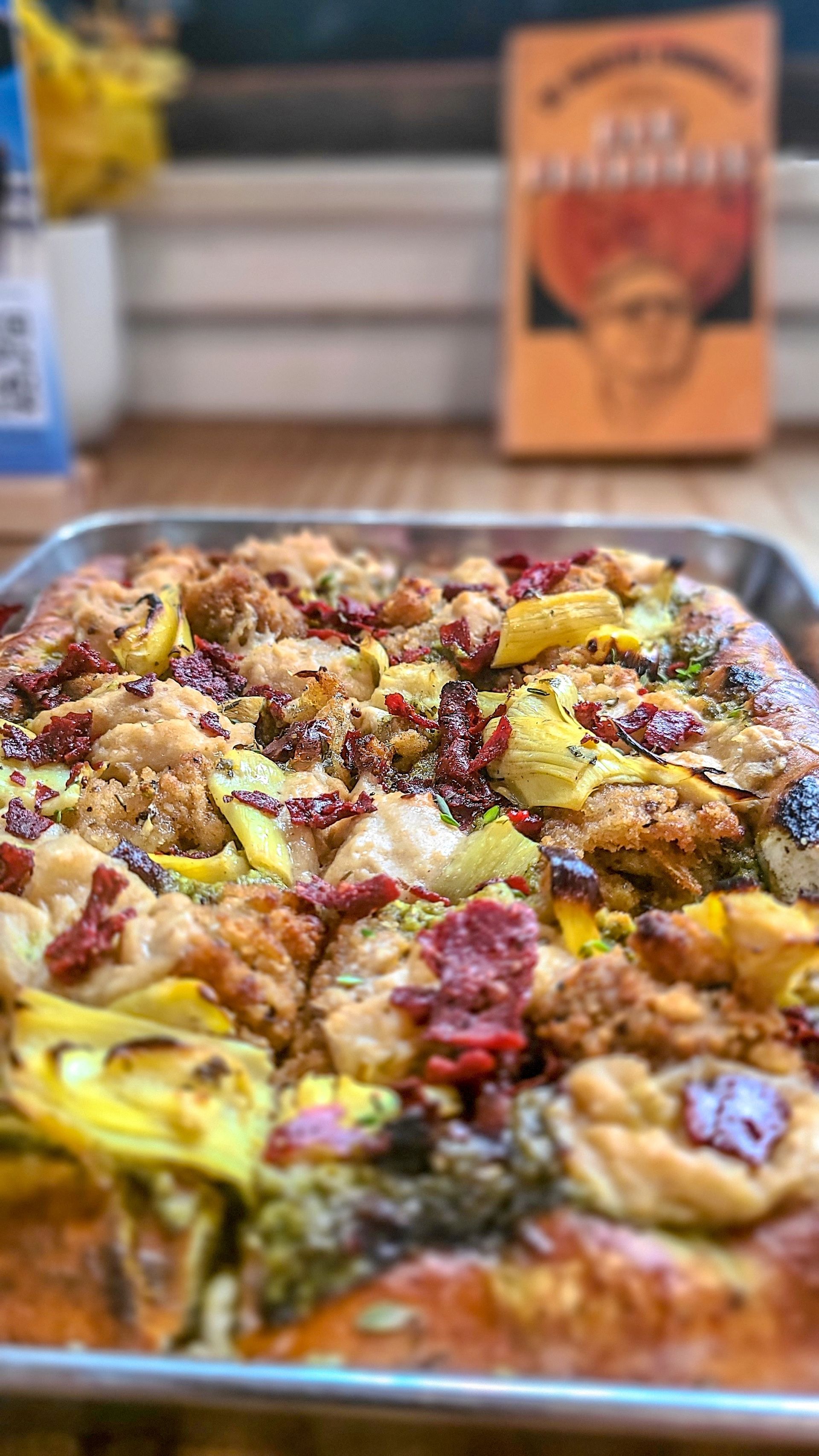 Close-up of a pan of focaccia bread topped with vegetables and meat, with a colorful menu in the background.