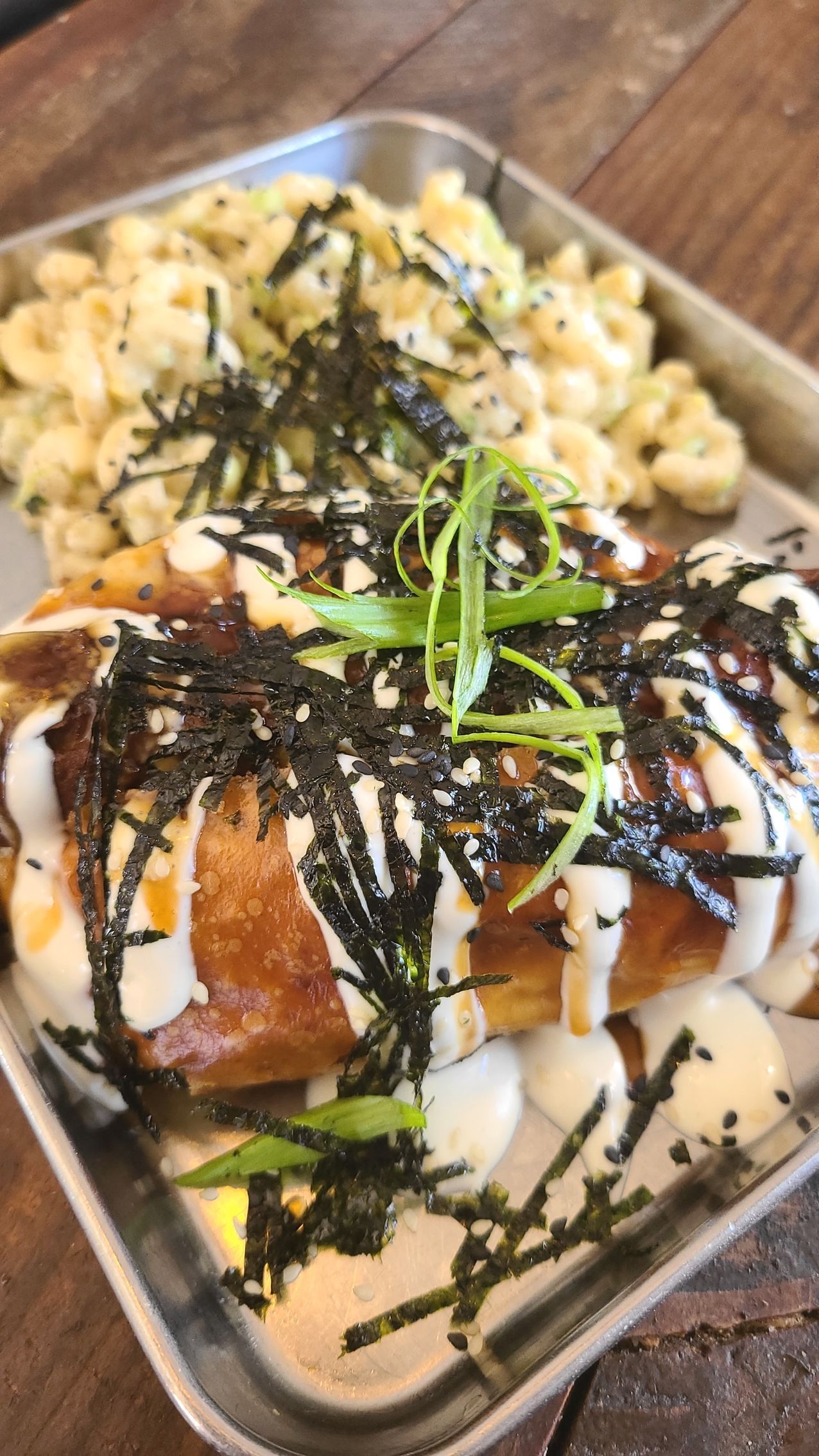 Hot dog topped with sauce and seaweed with mac and cheese on a metal tray.