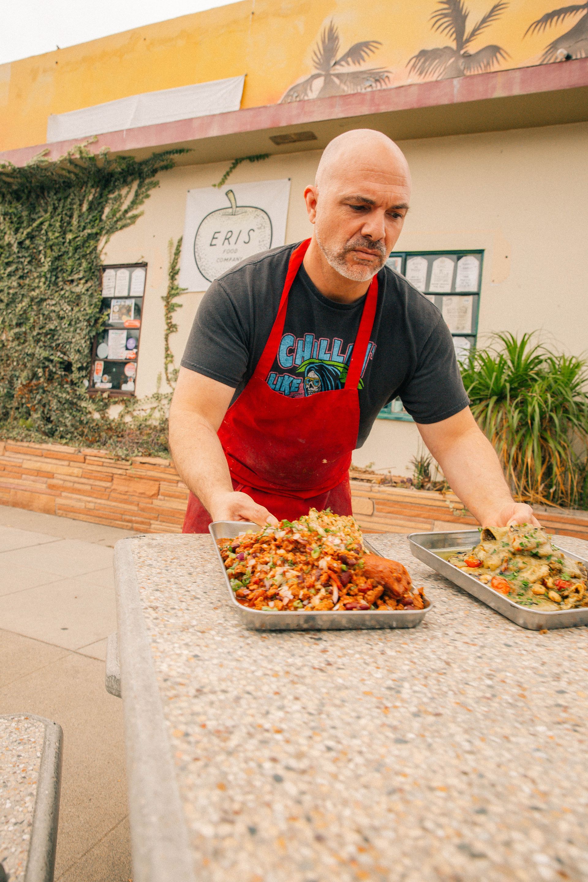 A man in a red apron prepares food on a table outside a restaurant.