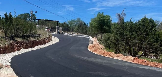 Asphalt road curves through a landscape of green trees, shrubs, and a clear blue sky.