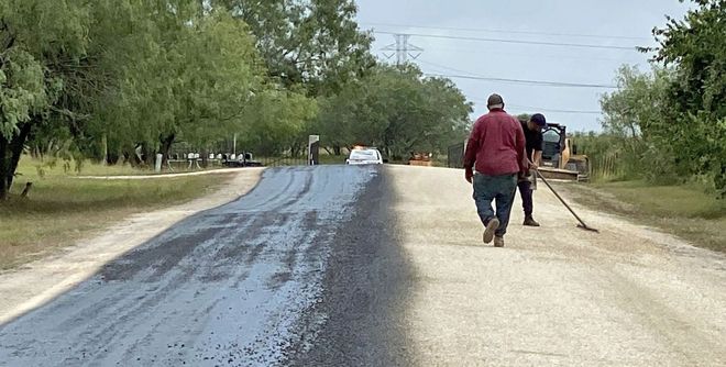 Workers paving a road. One spreads asphalt, other walks. Road half-paved, trees in the background.