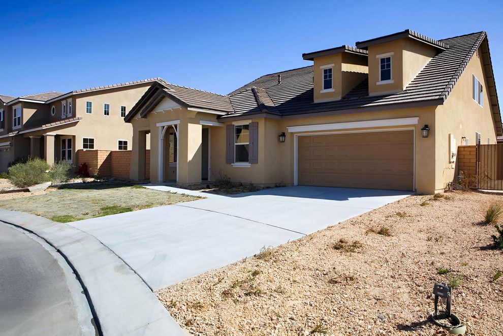 Tan house with a two-car garage, brown roof, and small front yard with gravel and sparse landscaping.