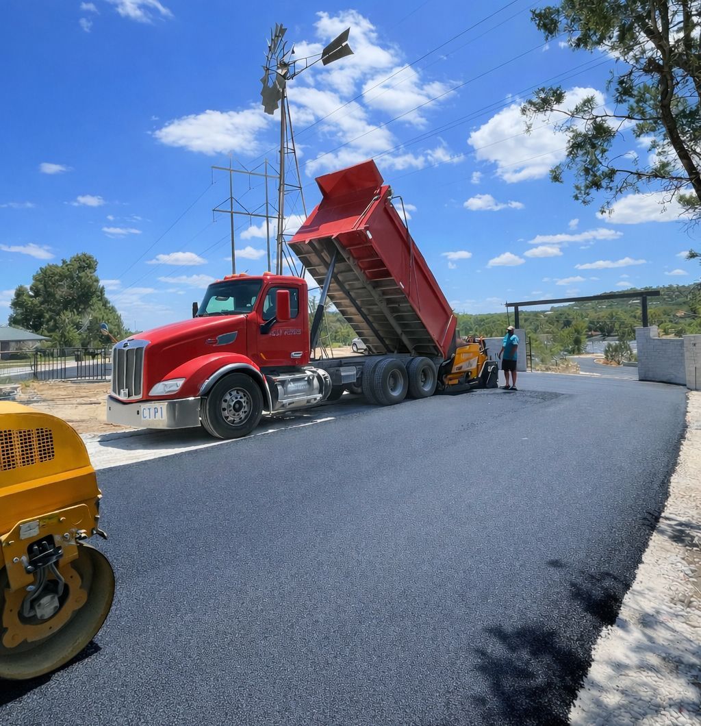 Red dump truck unloading asphalt onto a newly paved road; workers and a roller nearby.