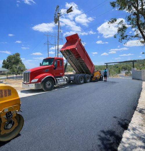 Red dump truck unloading asphalt onto a newly paved road; workers and a roller nearby.