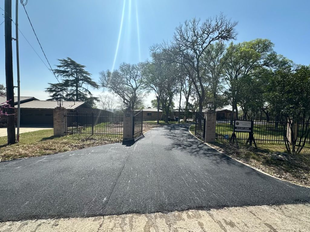 Paved driveway leading to a gated property with trees and a sunny sky.