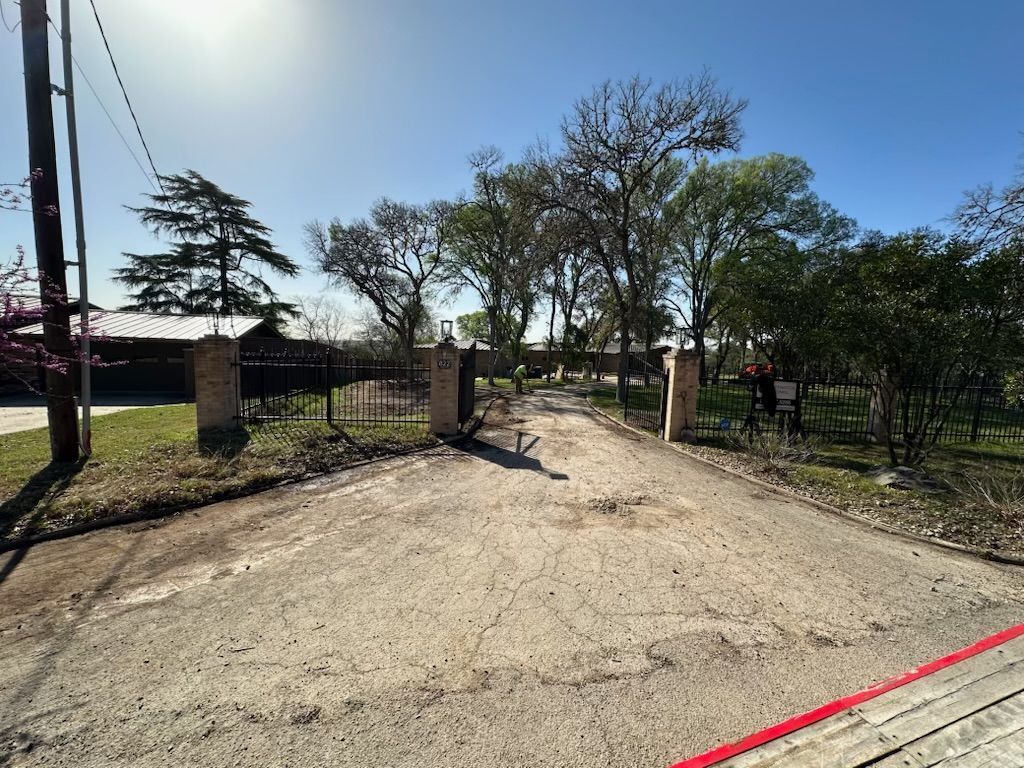 Entrance gate with black iron bars and stone pillars. Driveway leading towards trees and house on a sunny day.