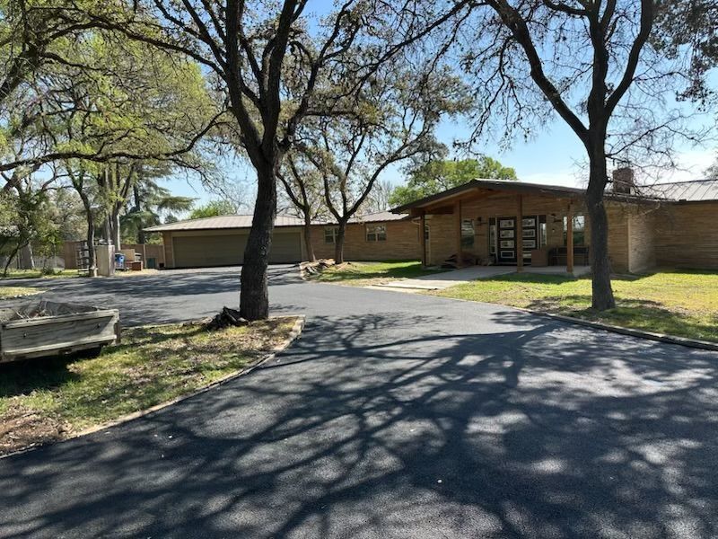 A one-story brown brick house with a dark asphalt driveway and shaded by trees.