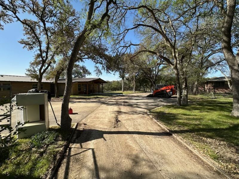A gravel driveway leads to a ranch-style home under trees. A small construction vehicle is in the distance.