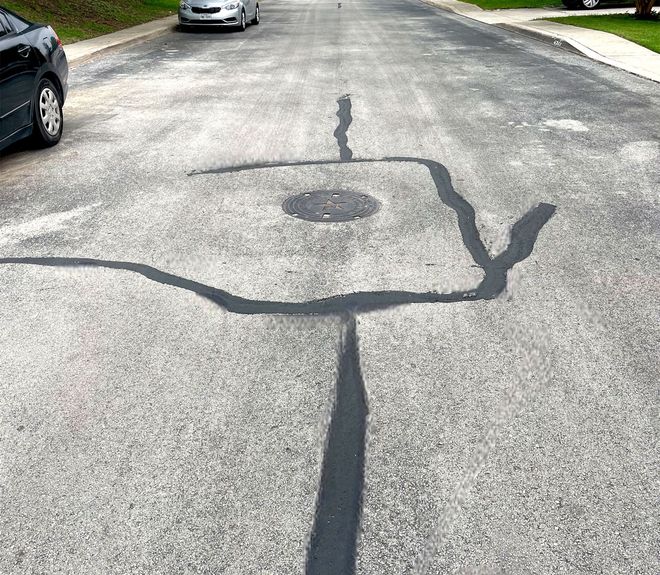 Asphalt road with dark sealant around a manhole cover and cracks. Cars are parked on the side.