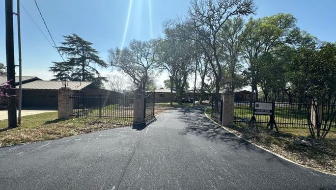 Paved driveway leading to a gated property with trees and buildings under a bright sky.