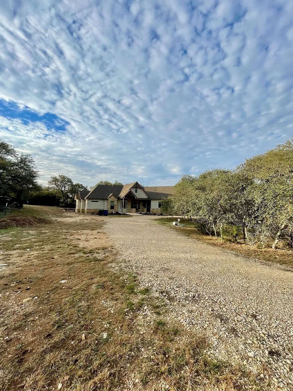 Gravel driveway leads to a one-story house under a cloudy sky. Trees line the right side.