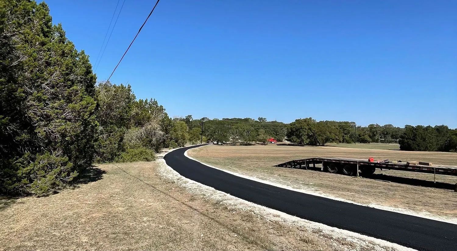 Paved path curves through a grassy field, bordered by trees on one side under a blue sky.