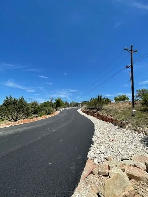 Paved road curves uphill, gravel shoulder on right, utility pole, blue sky, green scrub brush.