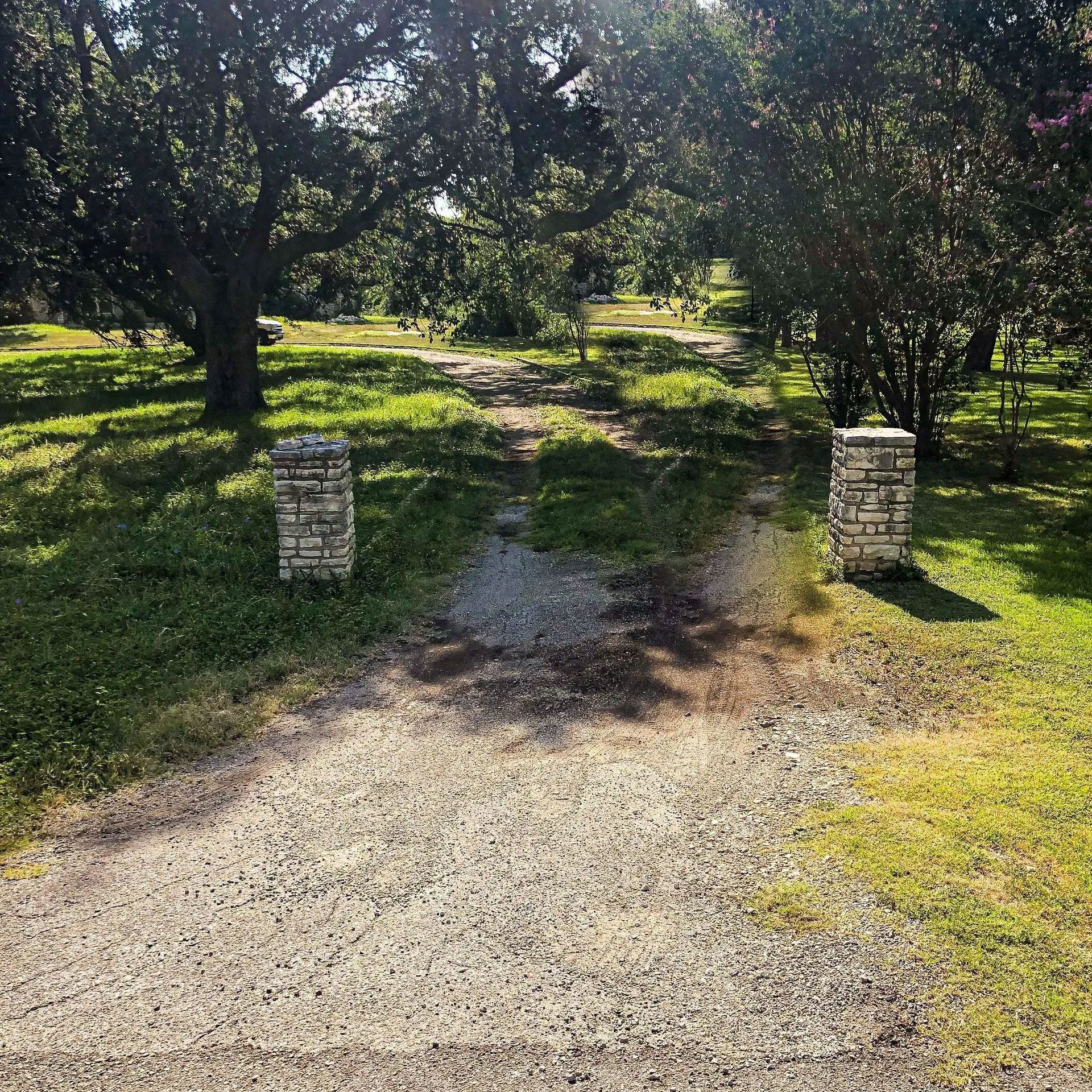 Gravel path leads between stone pillars, into a grassy yard shaded by trees.
