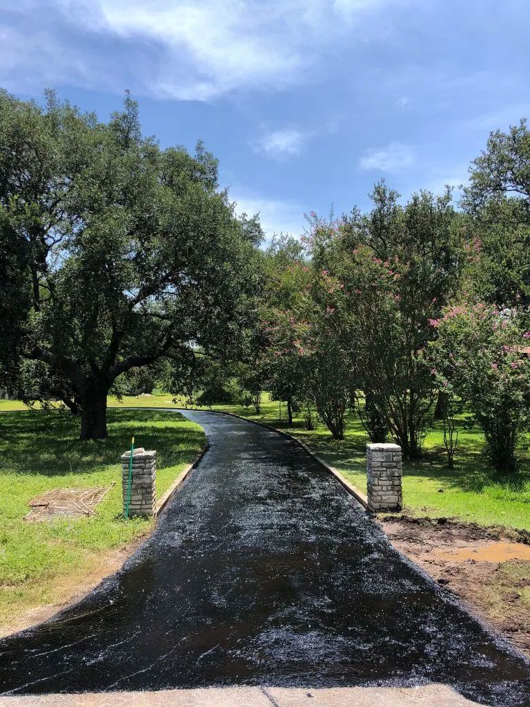 Freshly paved asphalt driveway leading toward trees under a blue sky.