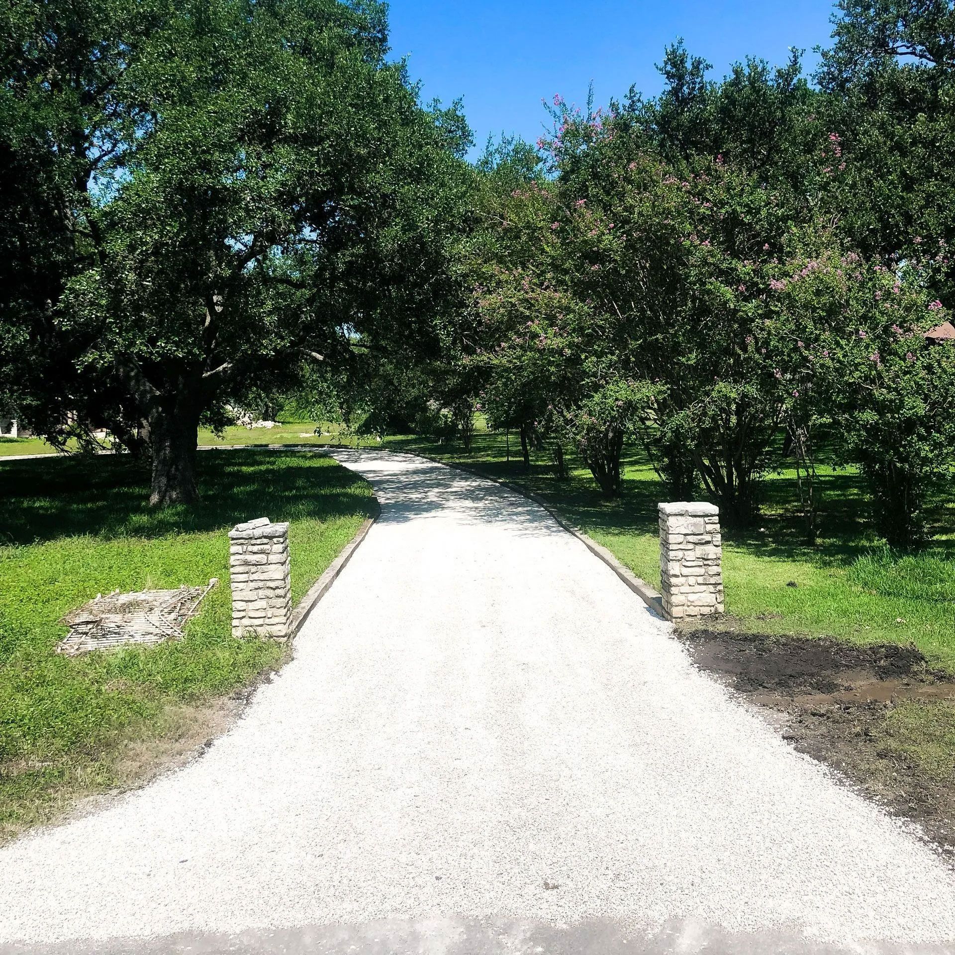 A white gravel driveway lined with brick pillars and trees, leading into a green, grassy area.