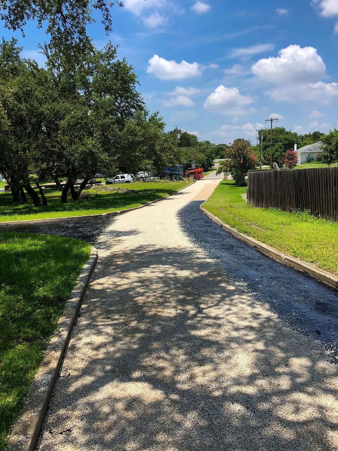 Gravel path lined with grass and trees, leading towards a street on a sunny day with blue sky and clouds.