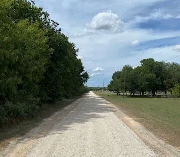 Gravel road through a rural area lined with trees and green grass under a partly cloudy sky.