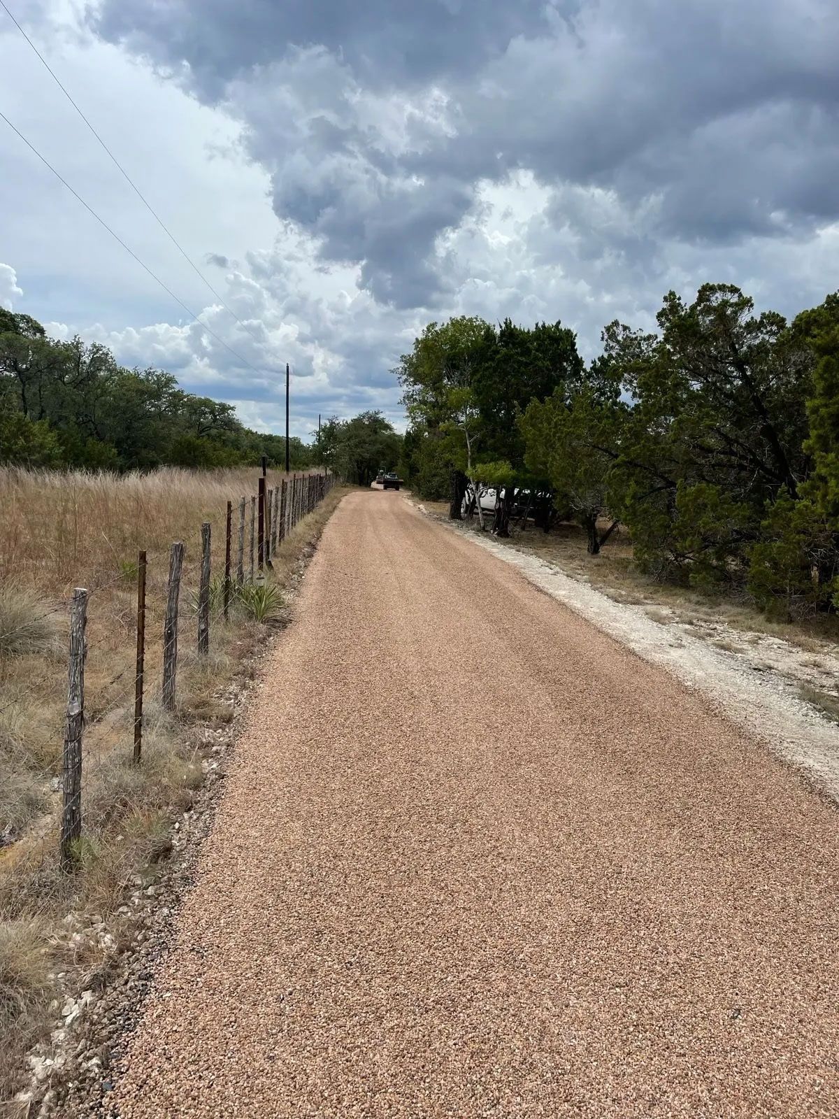 Gravel road through a rural area, lined by a fence and trees, overcast sky.