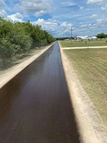 Freshly paved asphalt road running between dirt shoulders, trees, and grassy fields.