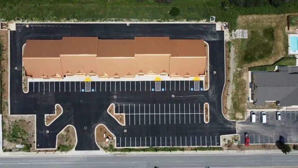 Aerial view of a building with a large parking lot, and a pool. Black asphalt, brown roof, and yellow parking space markers.