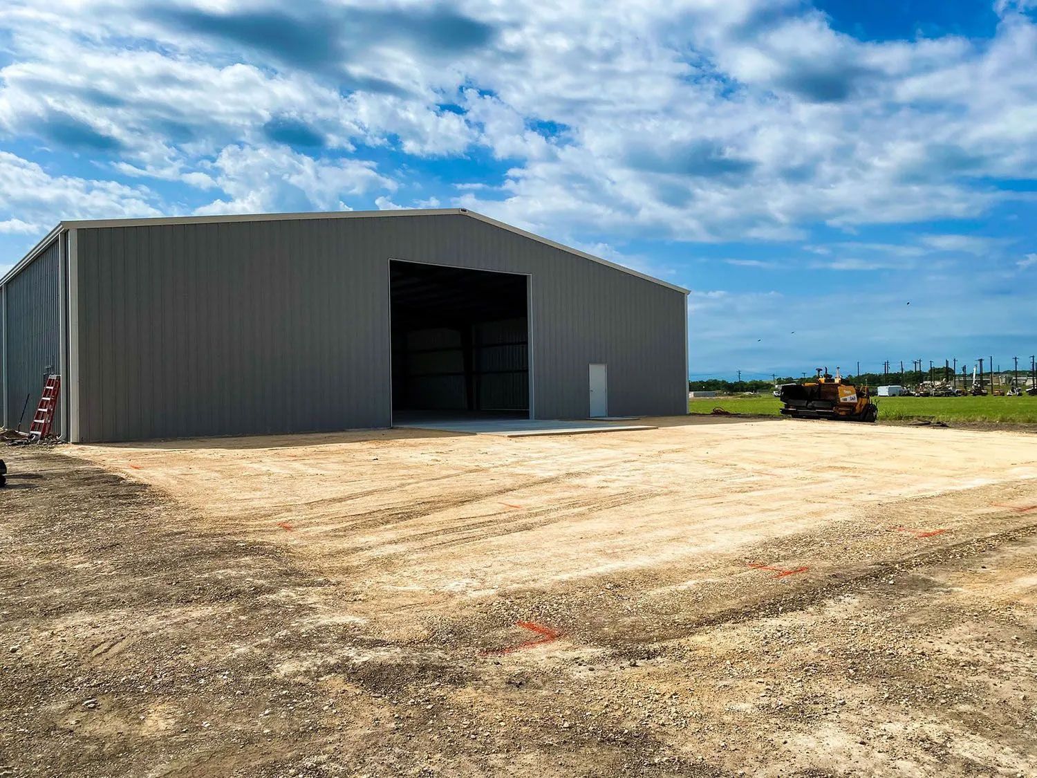 Gray metal warehouse with large open entrance on gravel lot under blue sky.