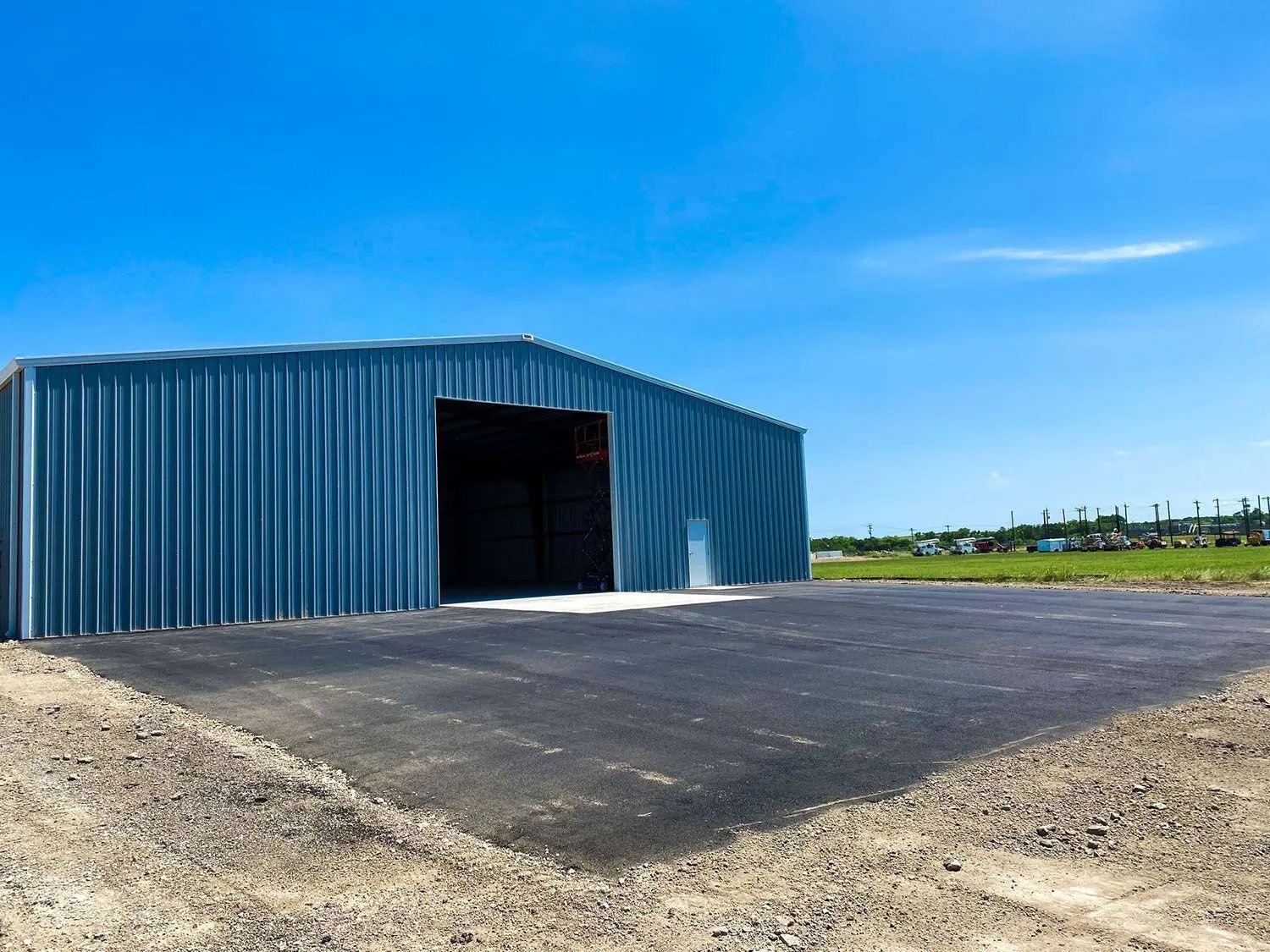 Blue metal shed with open black door, asphalt pad, gravel, and blue sky.