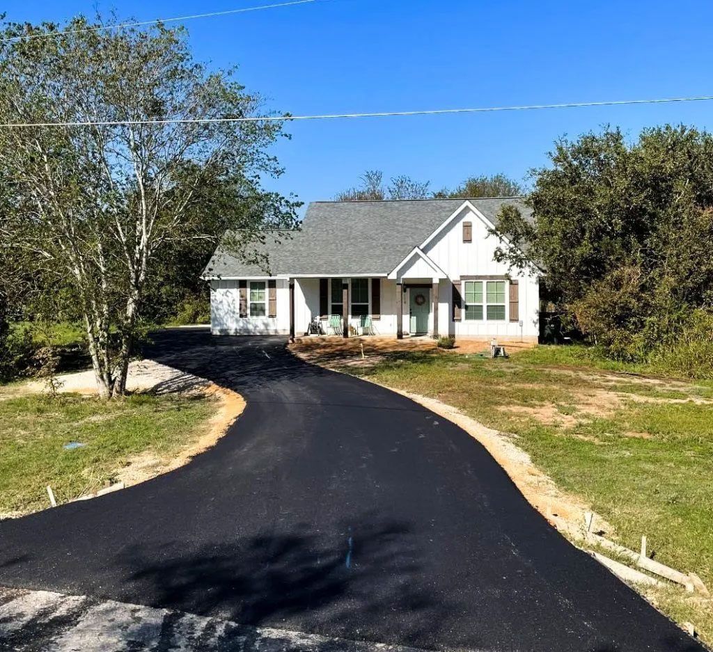 Asphalt driveway leading to a white house with brown shutters on a sunny day.