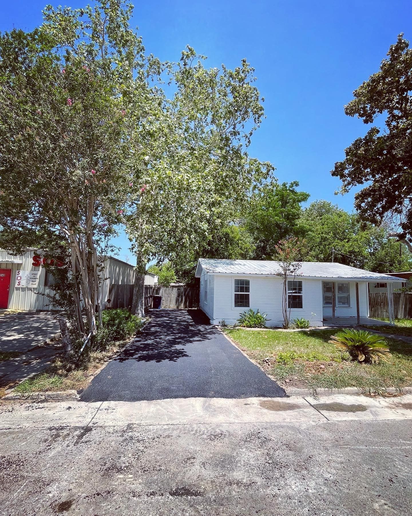White house with asphalt driveway under sunny blue sky, surrounded by trees.