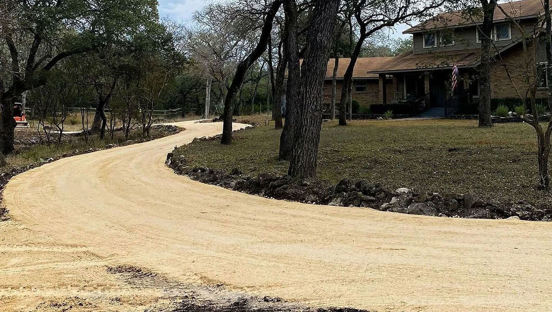 Winding beige gravel driveway leading to a two-story home, bordered by trees and lawn.