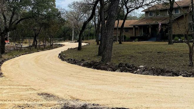 Winding beige gravel driveway leading to a two-story home, bordered by trees and lawn.