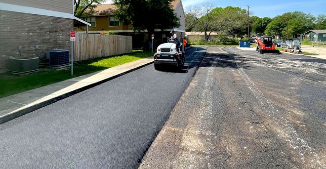 Road paving in progress. Newly paved road next to rough surface. Construction equipment visible.
