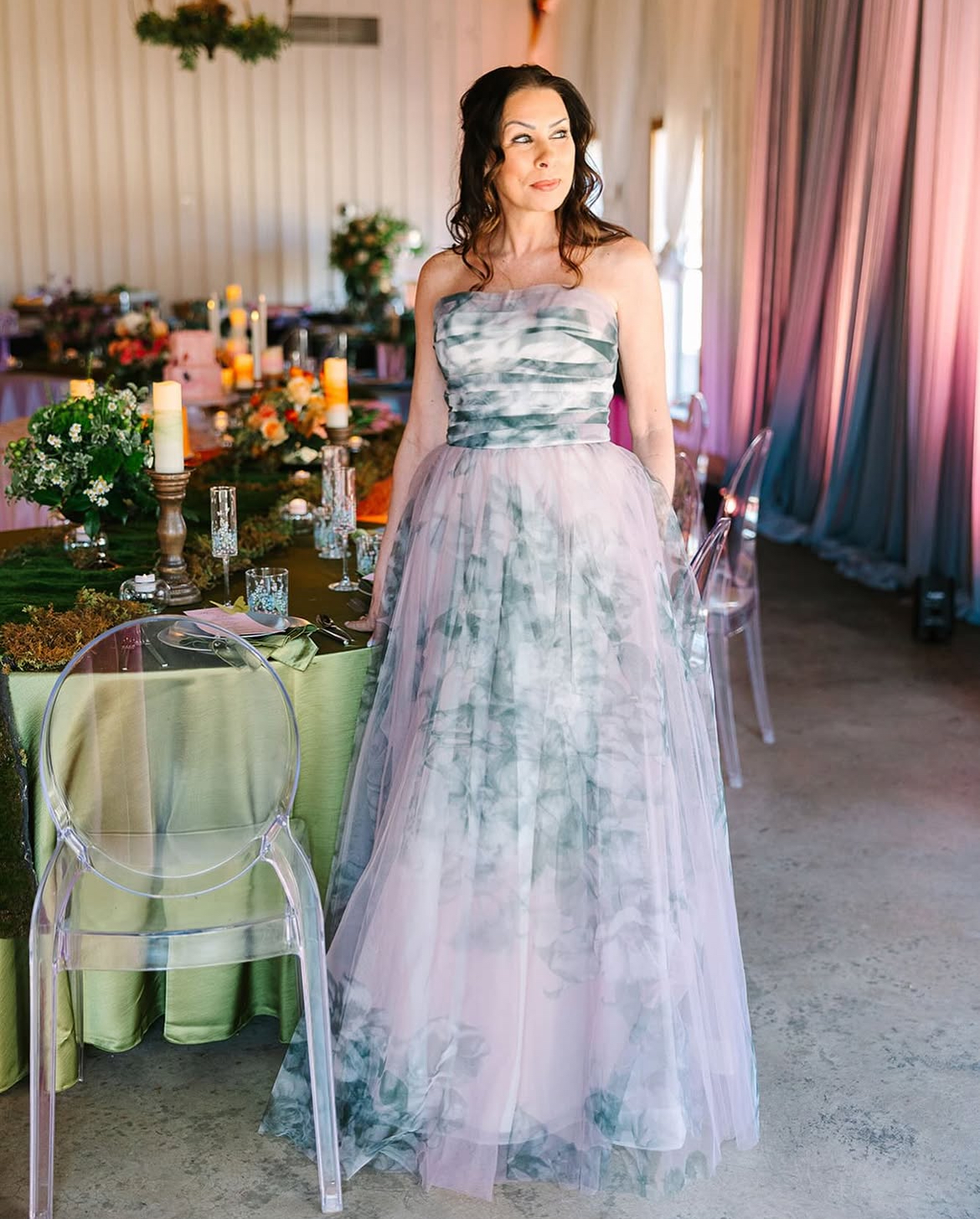 Woman in a strapless gown stands in a decorated room with a table, candles, and sheer curtains