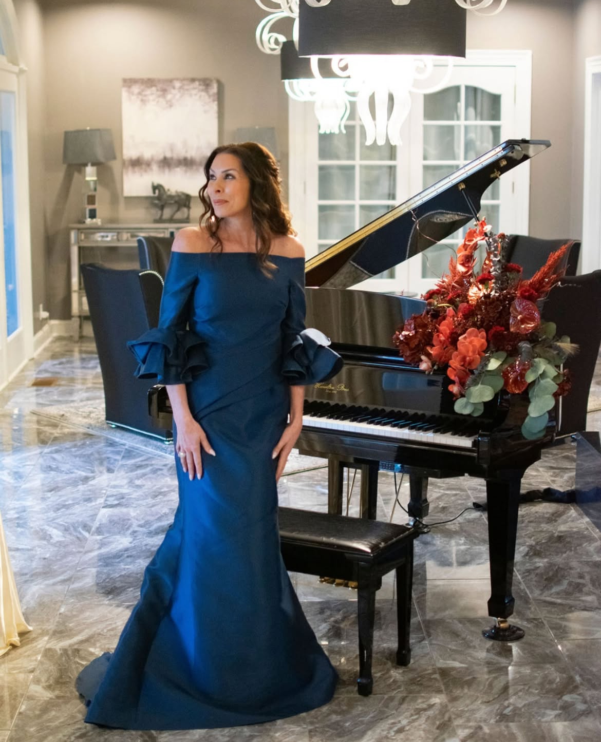 Woman in blue off-shoulder gown poses by a grand piano with a floral arrangement in a well-lit room