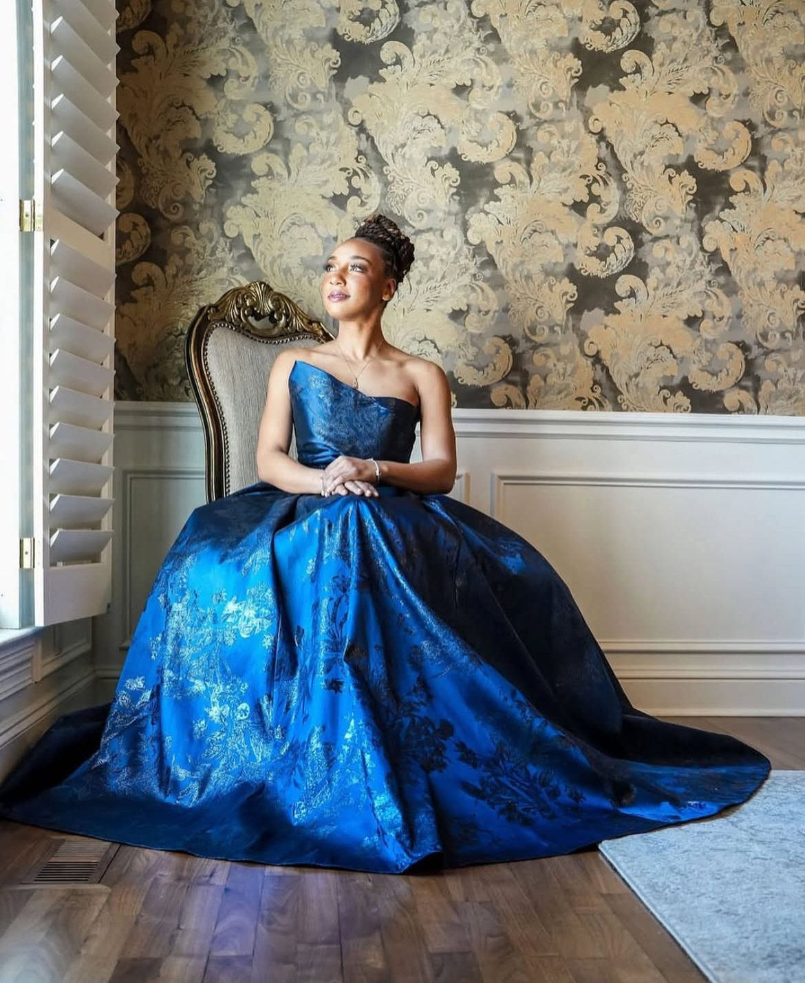 Woman in blue ball gown sits in a chair near a window with open shutters
