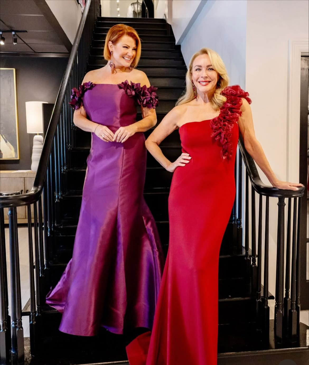Two women in formal gowns on a staircase. One in purple, the other in red, both with floral accents