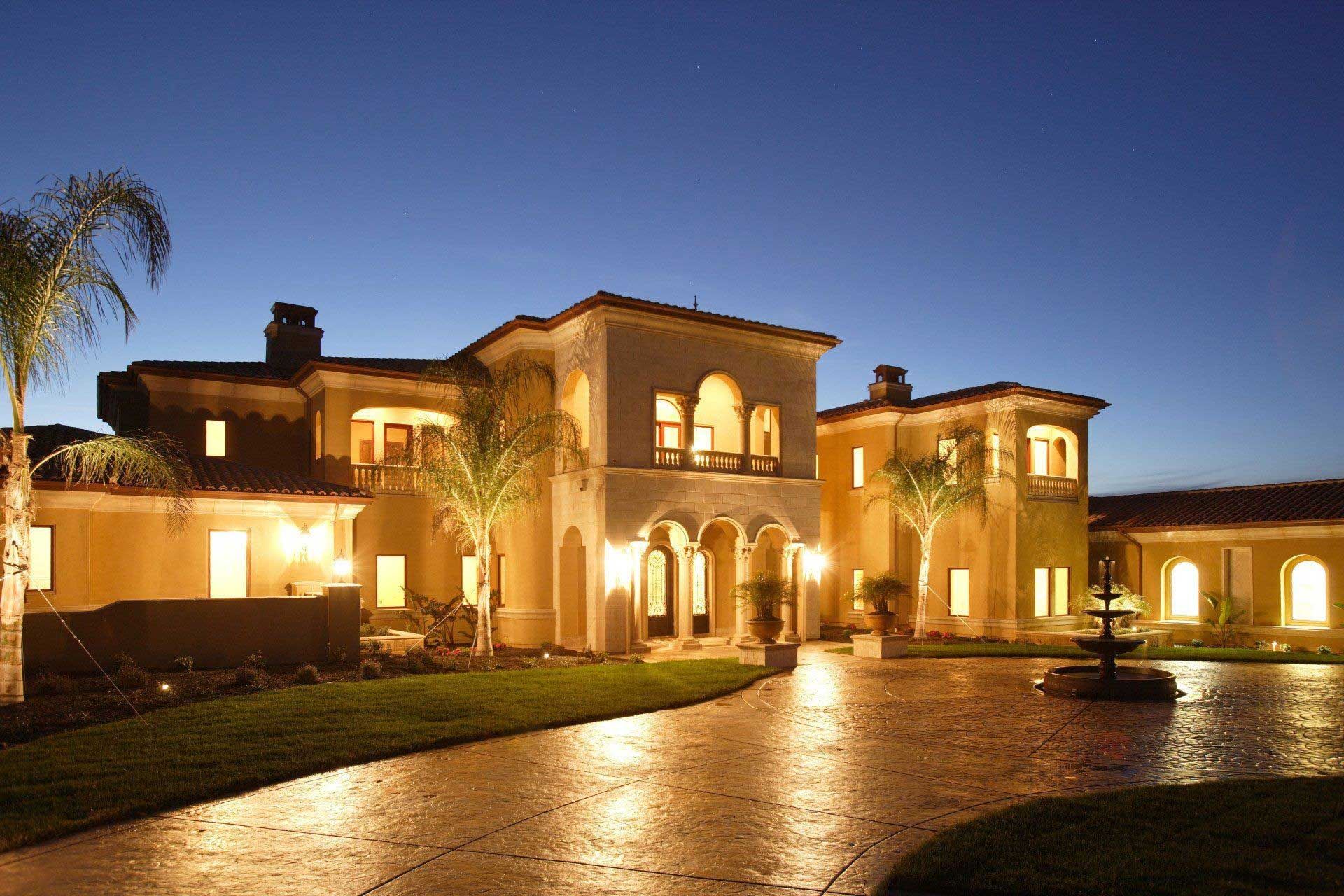Large, illuminated beige mansion with a circular driveway and fountain at twilight.