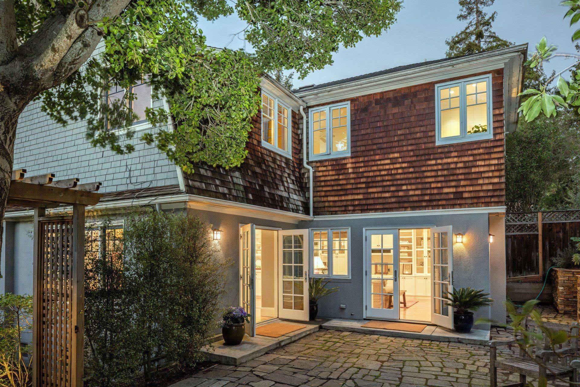 Back of a house with red shingles and a stone patio. Two-story with open French doors, light interior, and greenery.