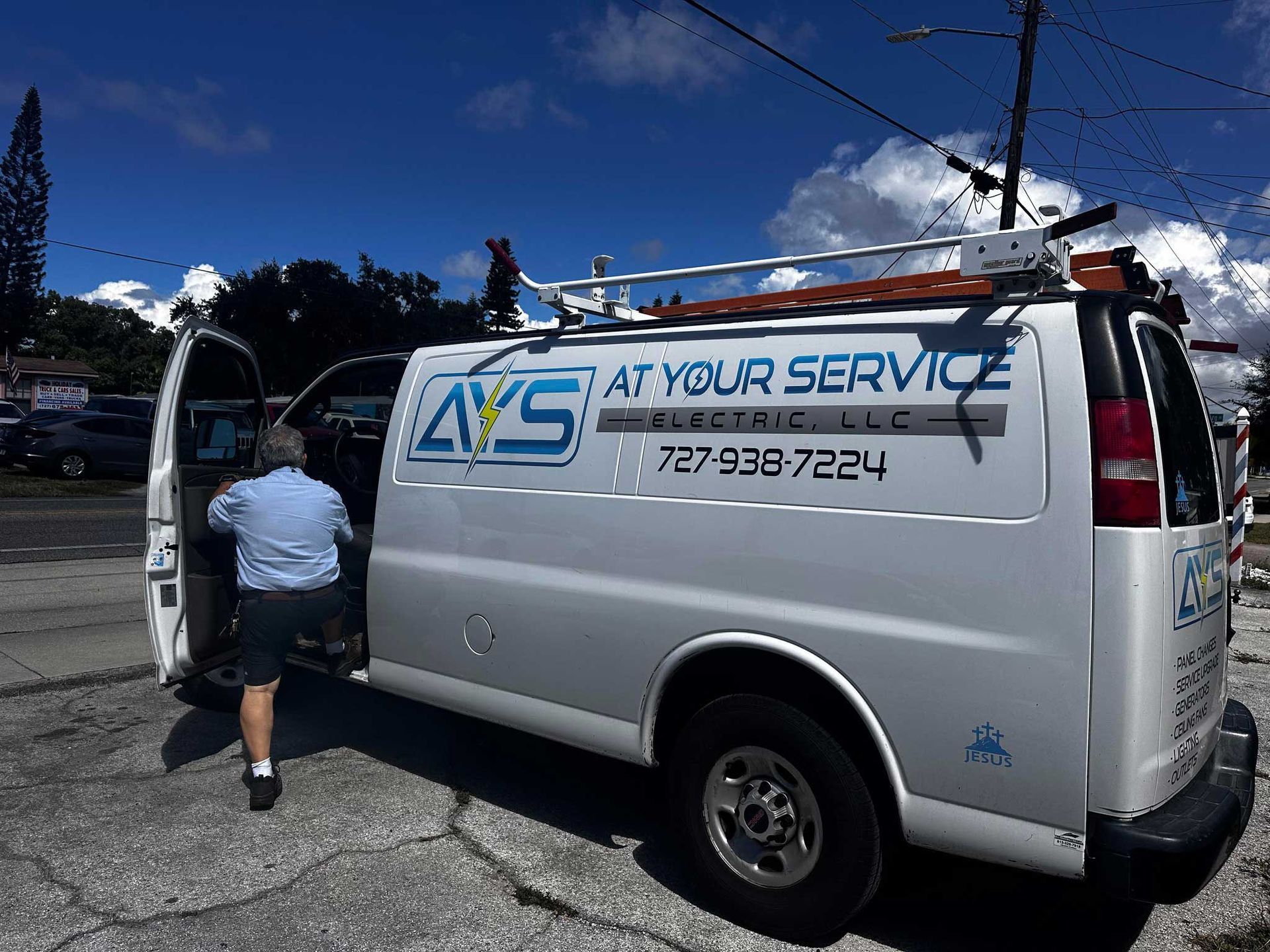 White service van with "At Your Service" logo, man entering the vehicle. Blue sky and parking lot in background.