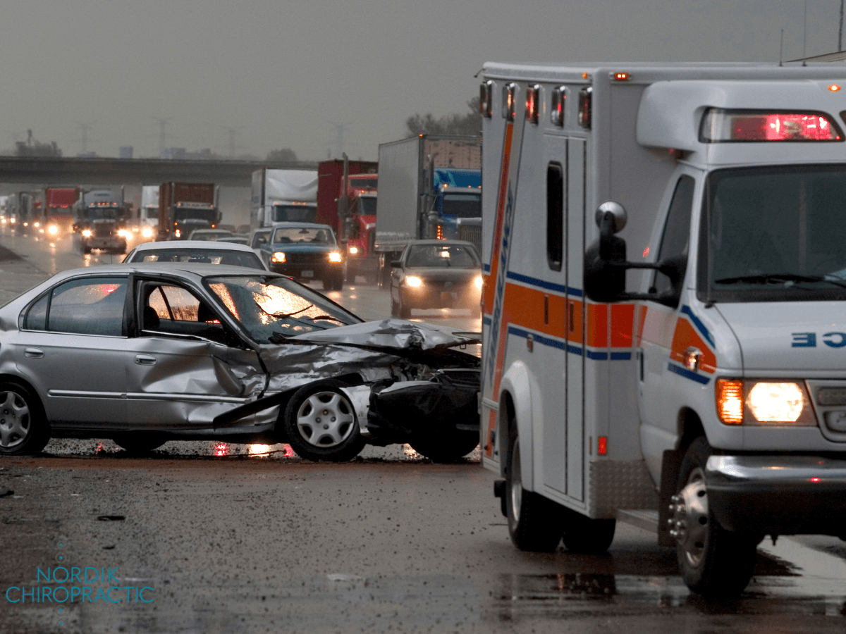 Damaged silver car in a multi-vehicle crash beside an ambulance on a wet highway